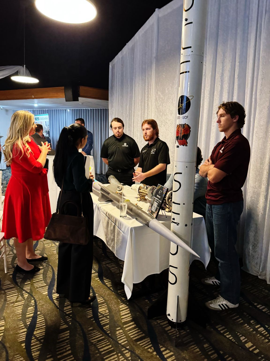 Attendees speaking with student exhibitors beside a UCF-branded rocket display